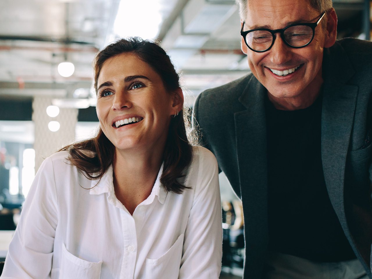 Two people smiling and leaning forward in a bright office, one wearing a white shirt and the other in a dark blazer with glasses.