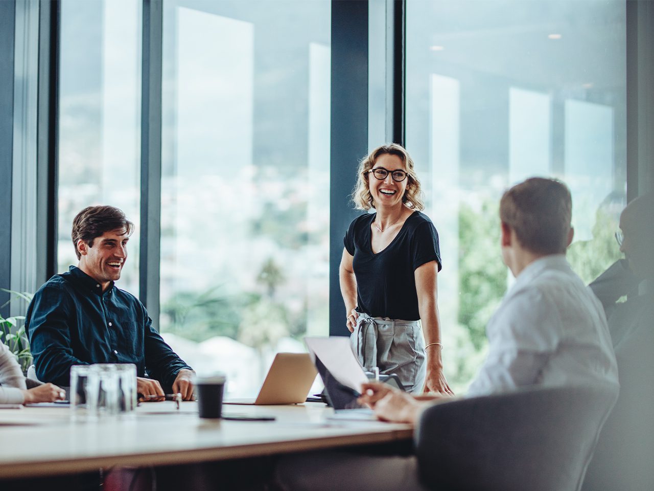 Lächelnde Person mit Brille steht am Konferenztisch in einem Büro, umgeben von Kolleg*innen, Laptop und Unterlagen; großes Fenster mit Stadtblick.