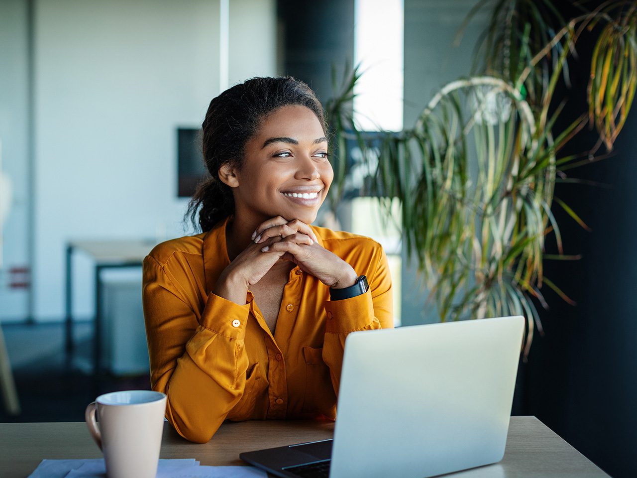 Una persona sorridente seduta davanti a un laptop, mani intrecciate sotto il mento, in ufficio con piante e una tazza.