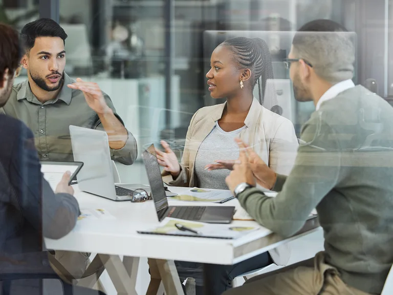 Diverse group of colleagues in a modern office, seated around a white table with laptops and documents, engaged in a discussion.