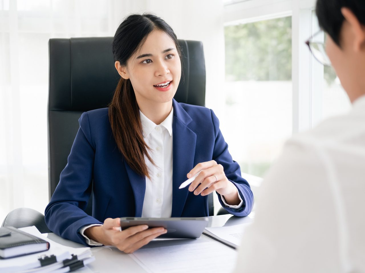A professional with long dark hair in a navy blazer sits at a bright office desk, holding a tablet and pen while talking to another person.