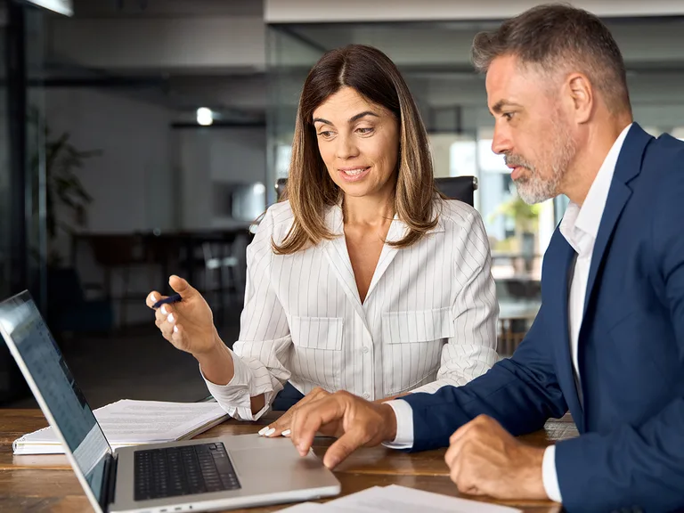 Two professionals in business attire sit at a wooden table with an open laptop and scattered papers, one holding a pen as they discuss.