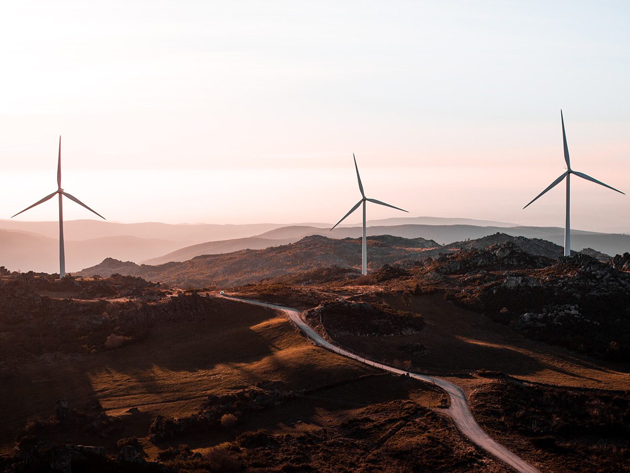 Three wind turbines on a hilly landscape at sunset, with a winding road through grassy terrain and distant mountain ranges in the background.