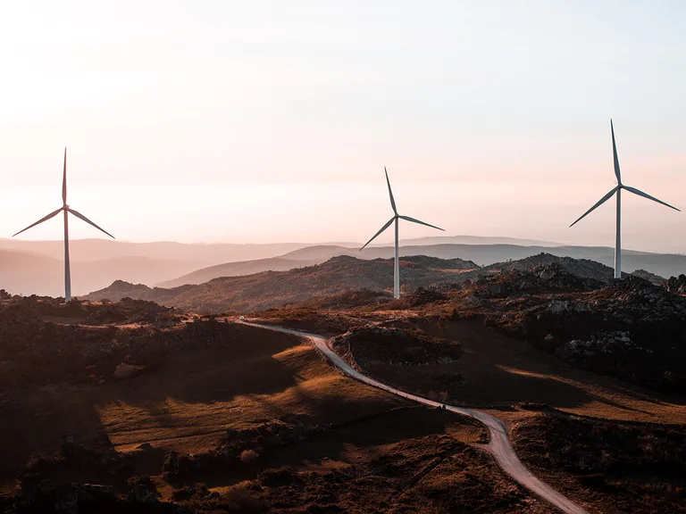 Three wind turbines on a hilly landscape at sunset, with a winding road through grassy terrain and distant mountain ranges in the background.