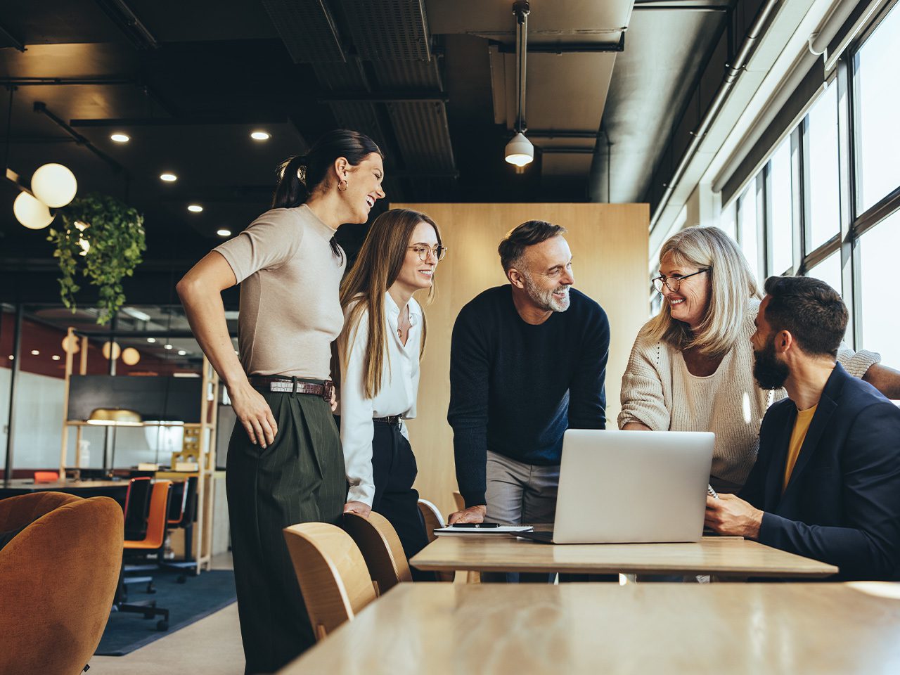 Five colleagues in a bright, modern office gather around a table with a laptop, smiling as they chat near large windows.