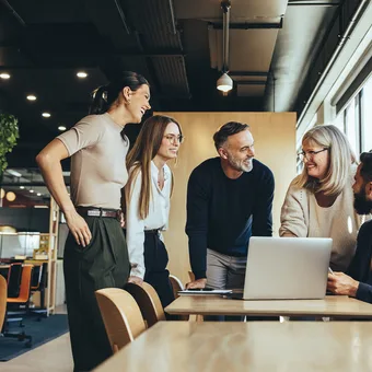 Five colleagues in a bright, modern office gather around a table with a laptop, smiling as they chat near large windows.