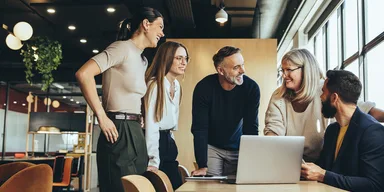 Five diverse colleagues gathered around a laptop on a wooden table in a bright, modern office, smiling and collaborating.