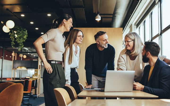 Five diverse colleagues gathered around a laptop on a wooden table in a bright, modern office, smiling and collaborating.