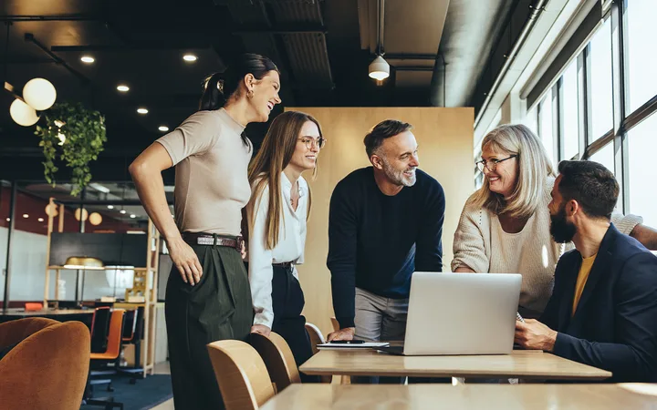 Five diverse colleagues gathered around a laptop on a wooden table in a bright, modern office, smiling and collaborating.