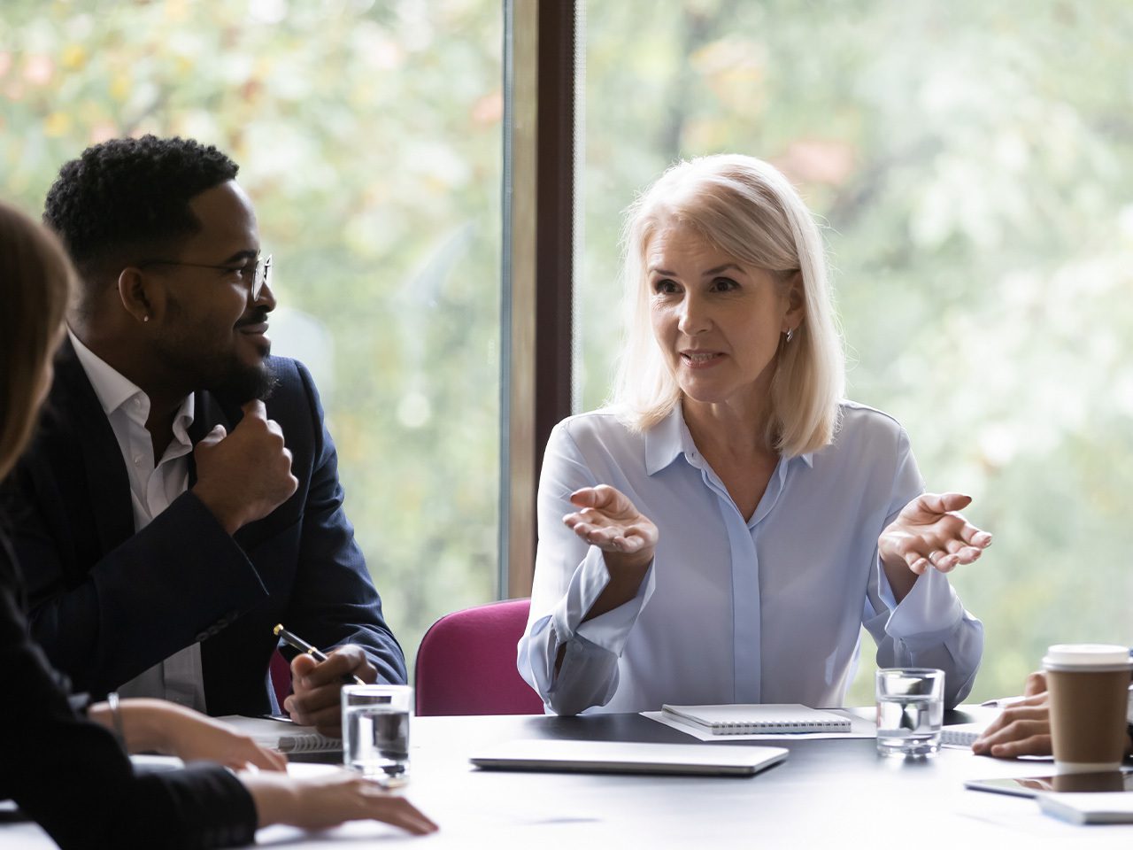 People at a meeting around a table by a window; a light-haired person in a pale blue shirt speaks with open hands, while others listen.
