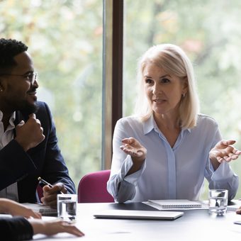 People at a meeting around a table by a window; a light-haired person in a pale blue shirt speaks with open hands, while others listen.
