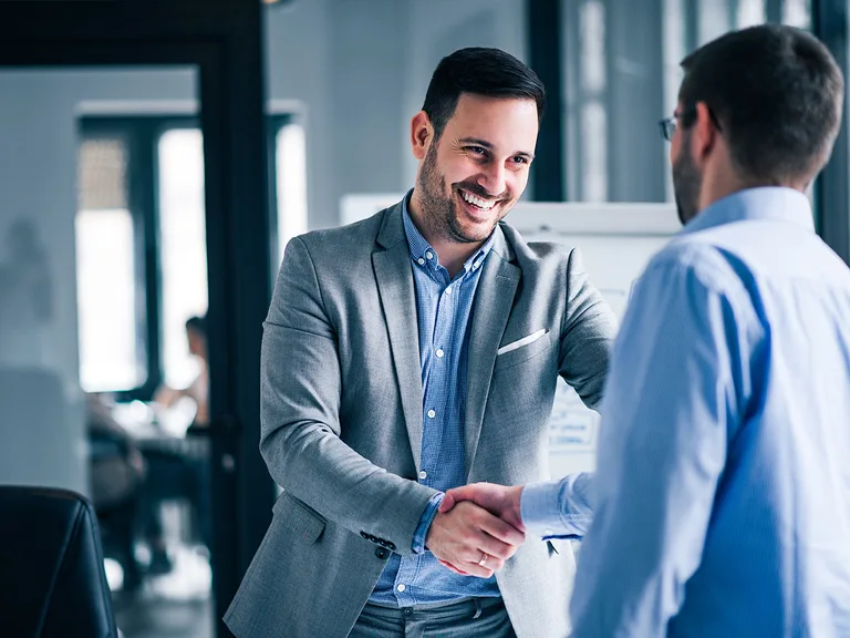 Two professionals shake hands in a modern office; one wearing a grey suit and blue shirt smiles, the other seen from behind with a blurred background.