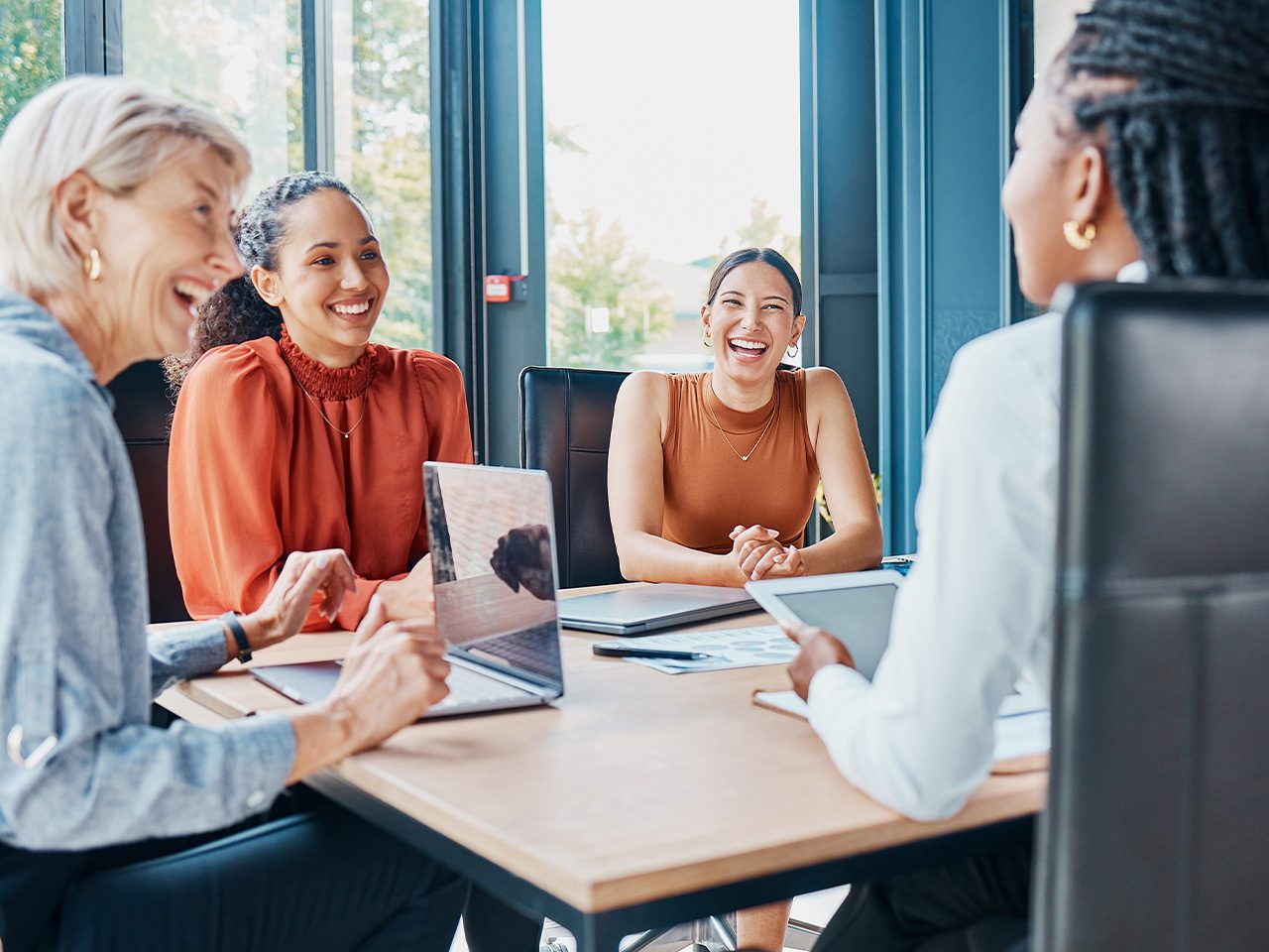 Four people sit around a wooden table in a bright office, smiling and chatting with laptops and tablets.