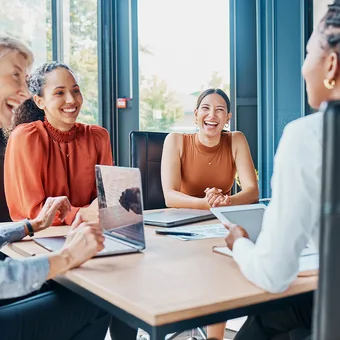 Four people sit around a wooden table in a bright office, smiling and chatting with laptops and tablets.