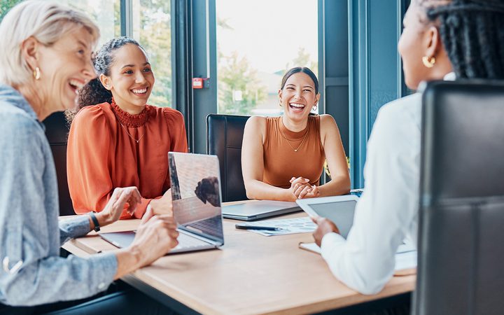 Four diverse people sit around a wooden conference table in a bright office, smiling and chatting with laptops and tablets.