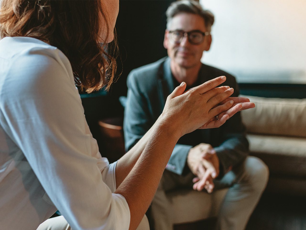 Two people in a living room; a person in a white shirt clasps their hands, while the other wearing glasses and a blazer sits on a sofa smiling.