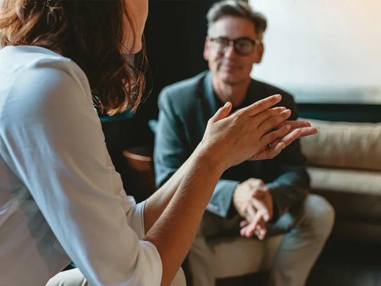 Two people in a living room; a person in a white shirt clasps their hands, while the other wearing glasses and a blazer sits on a sofa smiling.