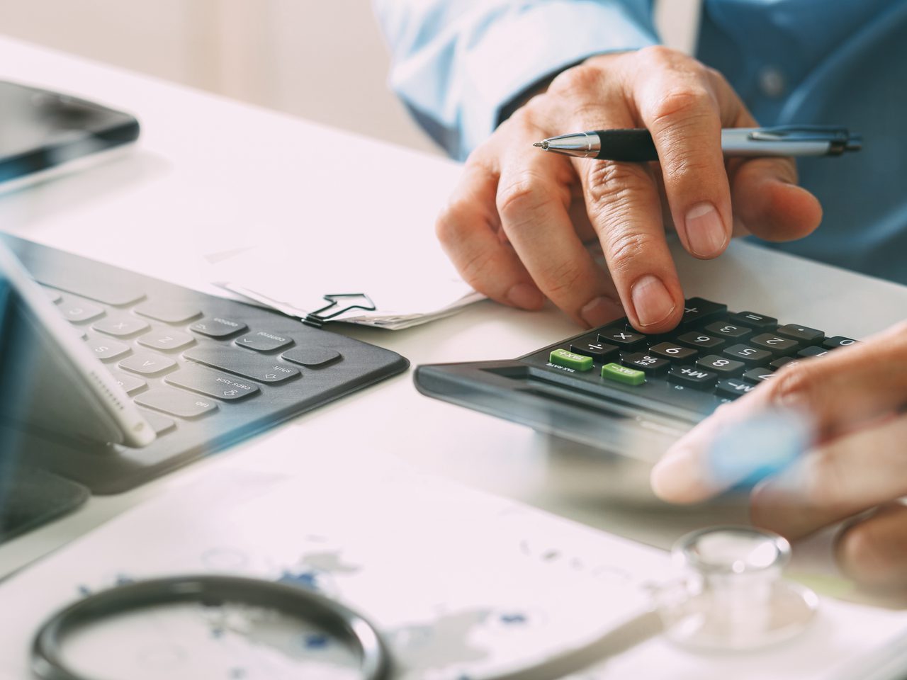 Close-up of a person’s hands using a calculator with a pen, on a desk with papers and a stethoscope nearby.