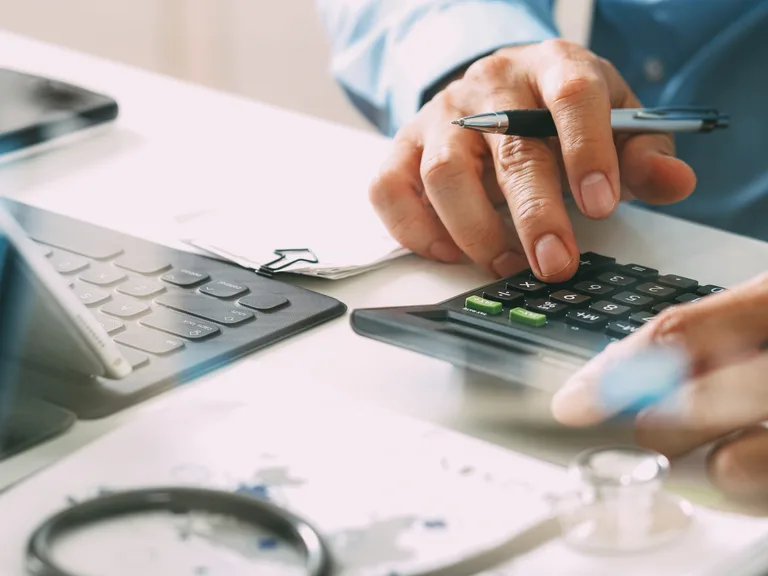 Close-up of a person’s hands using a calculator with a pen, on a desk with papers and a stethoscope nearby.