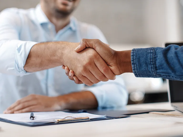 Two people shake hands over a desk with documents, a pen and a laptop, suggesting a business agreement.