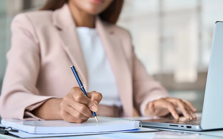 A person in a pale pink blazer writes in a notebook with a blue pen, beside a laptop on a bright office desk.
