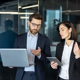 Two professionals in business attire discuss work in a modern glass-walled office, one with a laptop and the other with a tablet.