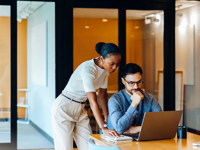 Two colleagues collaborate at a wooden table in a modern office; one stands and leans over a laptop while the other sits, focused on the screen.