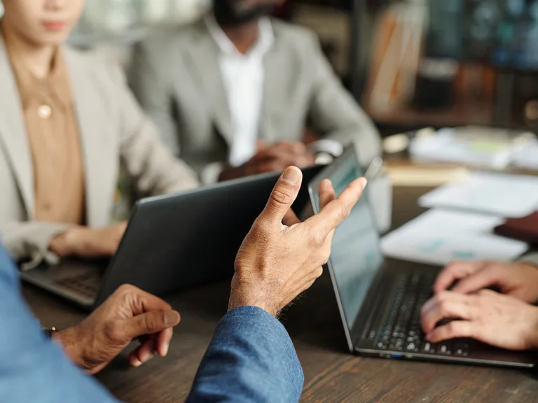 Hands hold a tablet at a wooden table as colleagues in blazers use laptops and review papers in a business meeting.