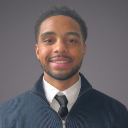 Person with short curly hair and beard, in a white shirt, black tie and navy jumper, smiling at the camera against a grey background.