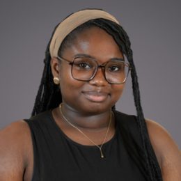 Portrait of a person with braided hair, wearing glasses and a beige headband, gold earrings and a pendant necklace, in a black sleeveless top.