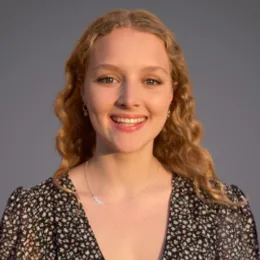 Portrait of a person with shoulder-length wavy blonde hair, smiling, wearing a black floral V-neck top, against a grey studio background.