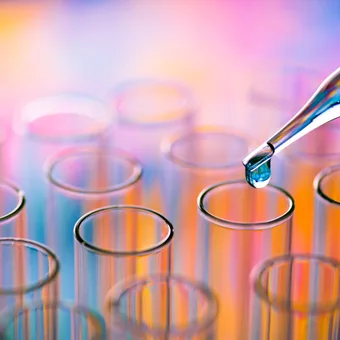 A close-up of a pipette releasing a droplet into a glass test tube, with other test tubes blurred in the colourful background.