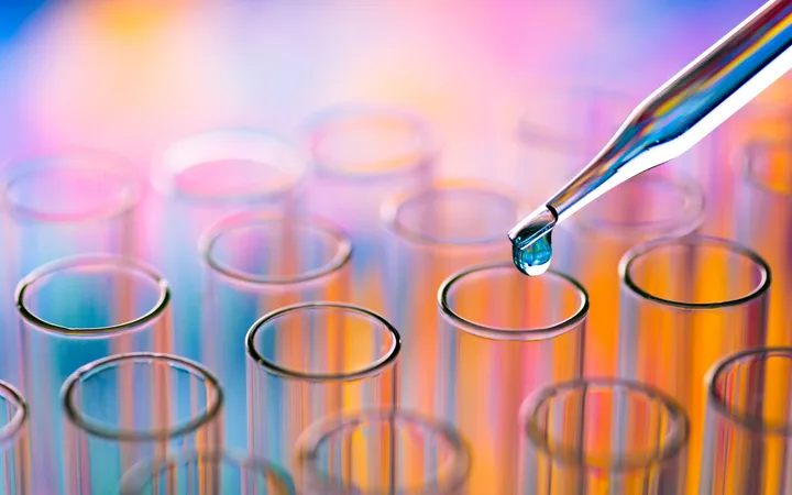 A close-up of a pipette releasing a droplet into a glass test tube, with other test tubes blurred in the colourful background.