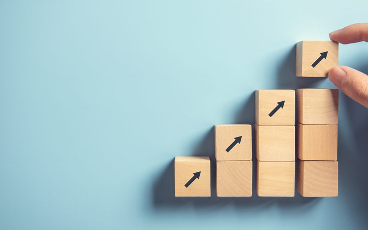 Hand placing a wooden block with an upward arrow on top of a series of stacked blocks, creating a rising bar chart on a blue background.
