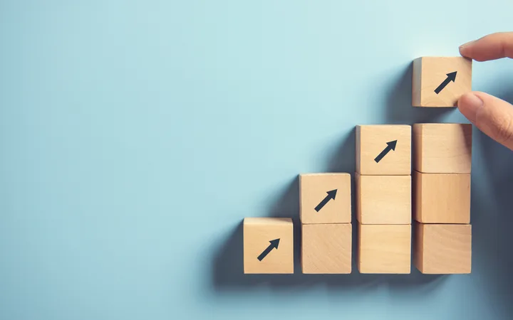 Hand arranging wooden blocks with upward arrows to form an increasing bar chart on a blue background.