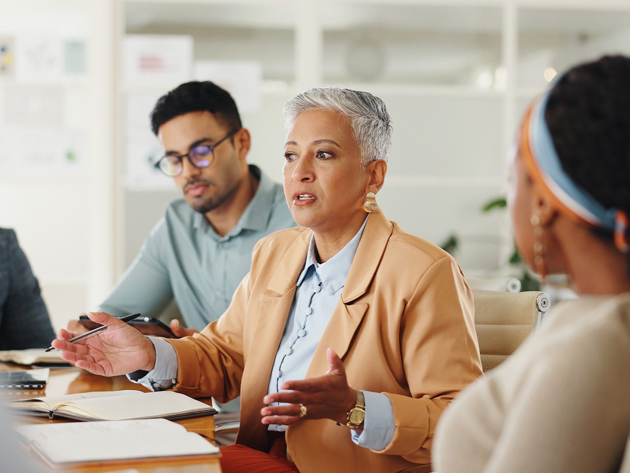 Personne aux cheveux gris courts en blazer beige et chemise bleu clair parle et gesticule lors d'une réunion de bureau; autres participants autour.