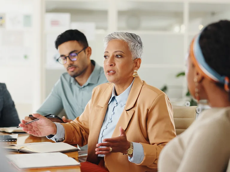 Personne aux cheveux gris courts en blazer beige et chemise bleu clair parle et gesticule lors d'une réunion de bureau; autres participants autour.