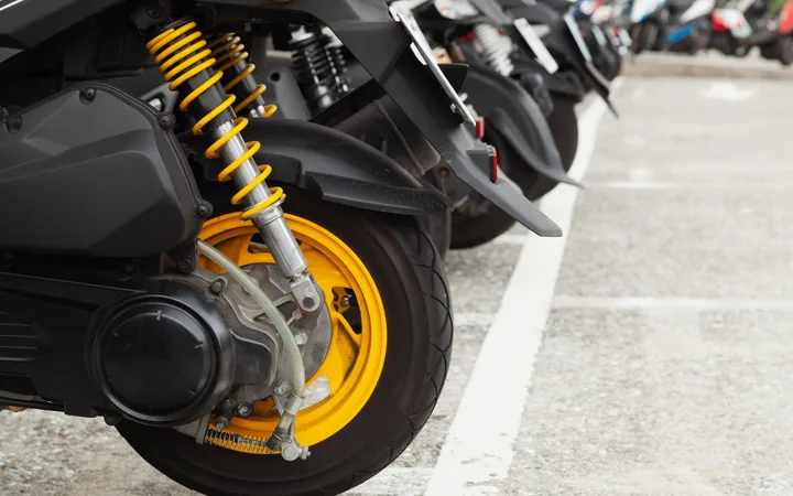 Close-up of a motorcycle’s rear wheel with bright yellow coil-spring suspension and yellow rim; other parked bikes blurred in the background.