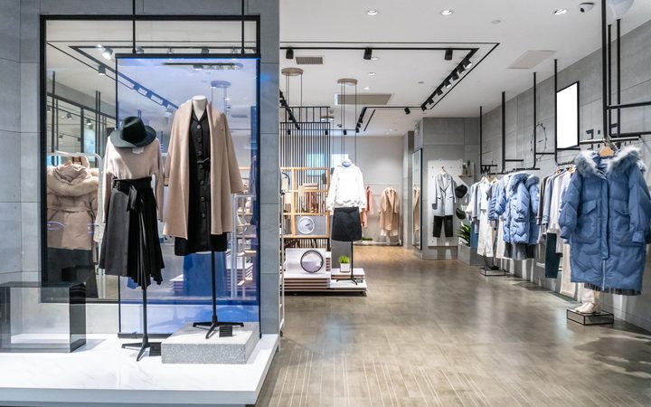 Modern clothing store with mannequins in beige and black outfits beside a glass display; racks of coats and jackets along the right wall, wooden floor.
