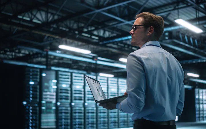 Person in a light blue shirt and glasses stands in a data centre, holding a laptop, with rows of illuminated server racks in cool blue lighting.