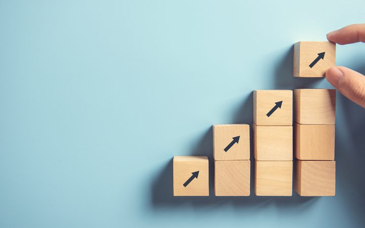Row of wooden blocks stacked as an ascending staircase with upward arrows; a hand places the top block on a blue background.