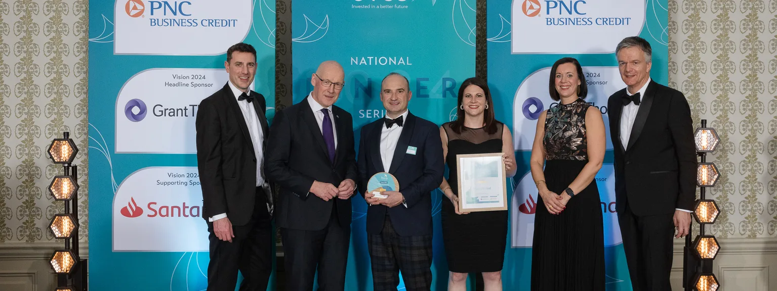 Six people standing on a stage with blue banners in the background, dressed in formal attire, posing for a photo at an awards event.