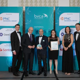 Group of six people dressed in formal attire standing on a stage with blue banners in the background at a corporate event.