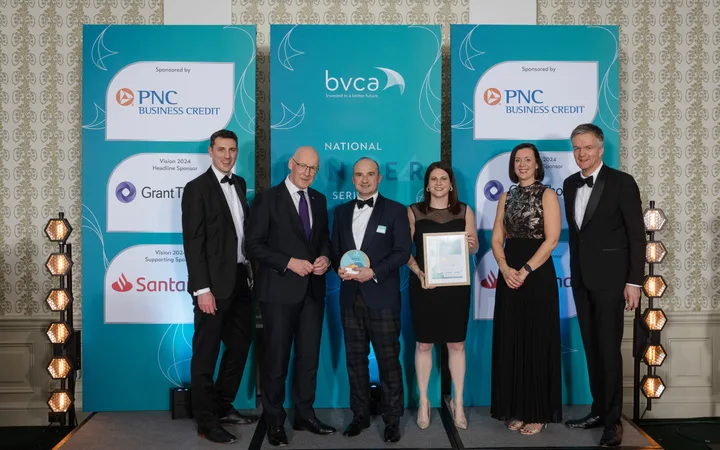 Group of six people dressed in formal attire standing on a stage with blue banners in the background at a corporate event.