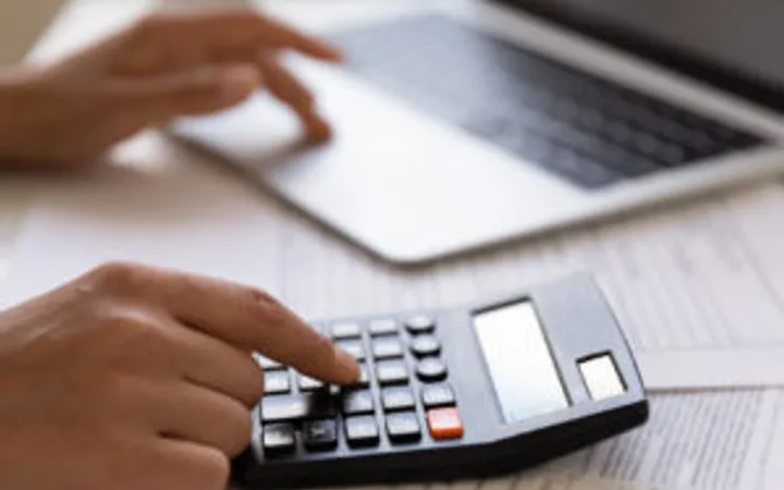 A person's hand using a calculator on a desk, with an open laptop, papers, and a plant in the background.
