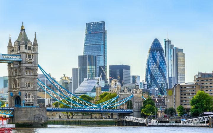 Tower Bridge spans the River Thames in front of London's modern skyline, including The Gherkin and other glass skyscrapers under a clear blue sky.