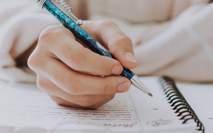 A person writes in a spiral notebook with a blue and black pen, focusing on their hand and the notebook, with a blurred background.