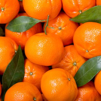 Close-up of a pile of bright oranges with glossy peels, a few attached stems, and scattered green leaves.