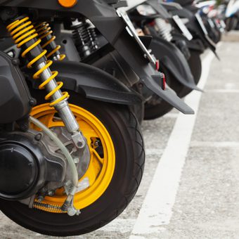 Close-up of a black motorcycle rear wheel with bright yellow suspension springs and yellow rim, parked among other bikes in a paved car park.