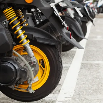 Close-up of a black motorcycle rear wheel with bright yellow suspension springs and yellow rim, parked among other bikes in a paved car park.